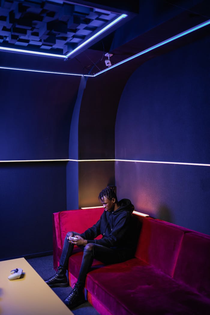 Young adult gaming on a red sofa in a neon-lit room.