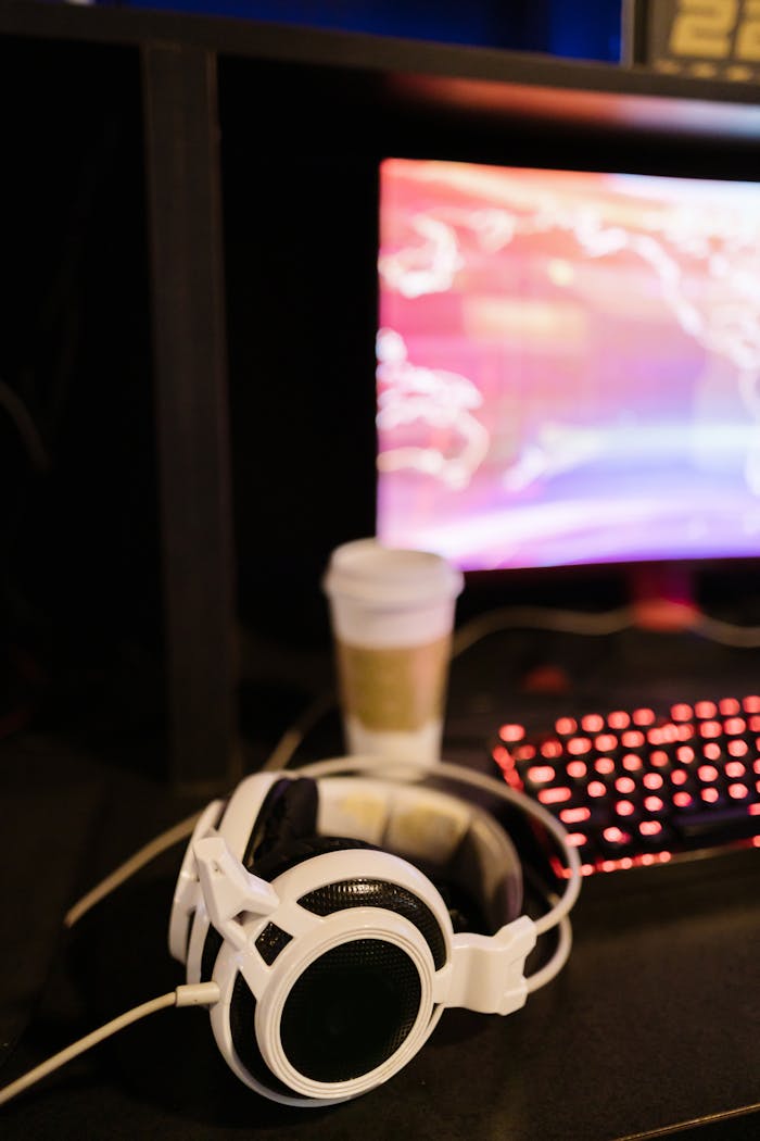 A gaming desk setup with white headphones, keyboard, blurred screen, and coffee cup.